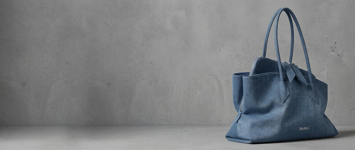 The Attico La Passeggiata medium tote bag in washed blue denim against a minimalist cool grey concrete background from The Exclusive Line.