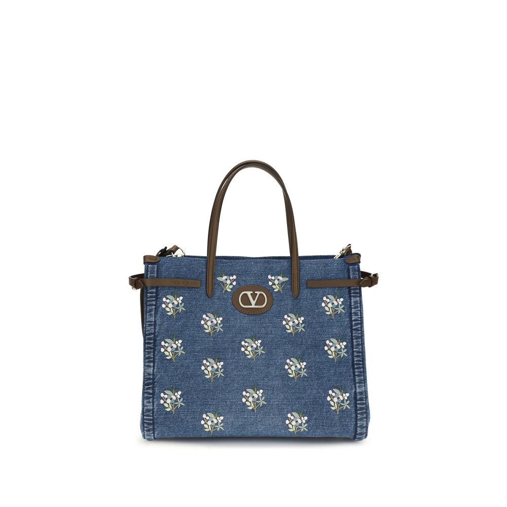 Valentino Garavani Antibes medium shopping bag in blue denim featuring white floral embroidery, leather top handles, and gold VLogo hardware details.